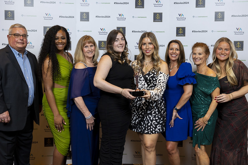 A group of individuals in formal attire pose for a photo in front of a logo wall, while the two in the center hold an award