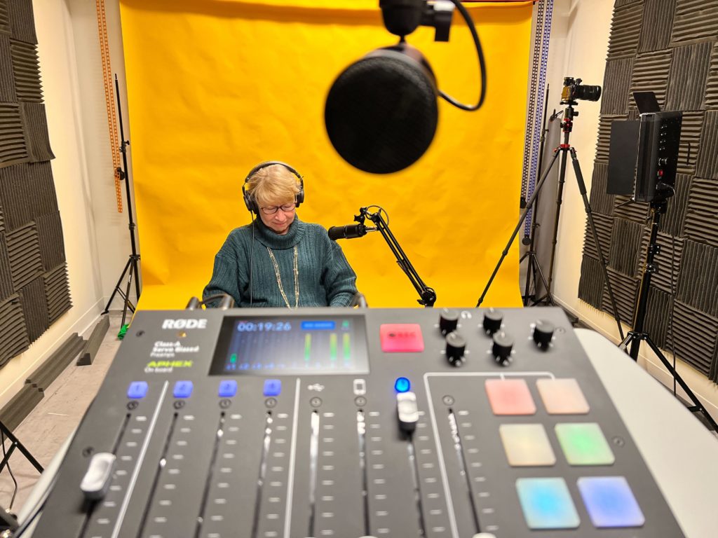 Candee Chambers in the DE Talk studio sitting behind an audio control panel and microphone, in front of a yellow backdrop.