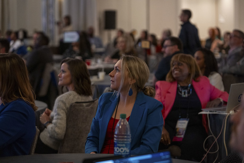 Women sit at tables during a General Session with a sea of attendees in the background.