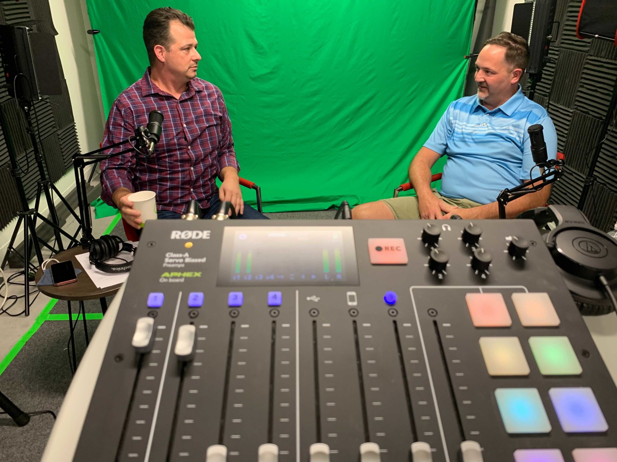 In the foreground sits a Rode podcast recorder, with guests Jason Ward and Scott Sendelweck in front of a green screen. Jason is white male in his 40's with brown hair, wearing a plaid button down shirt. Scott Sendelweck is also a white male in his 40's with brown hair and a goatee, wearing a blue striped polo and khaki shorts.