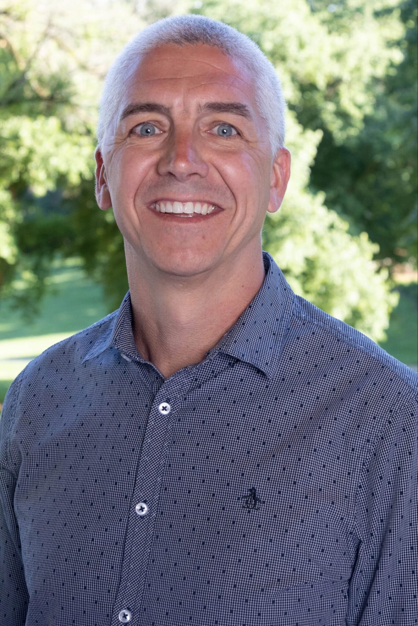 Professional headshot of a bald, white man smiling against a green tree backdrop, wearing a blue collared button up shirt; Name: Josh Christianson, PEAT