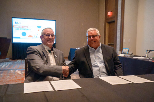 Two men in business attire are seated at a conference table, smiling and shaking hands. In front of them are several official documents, suggesting a formal agreement or partnership. Behind them, a presentation screen displays the NLx logo and the phrase 