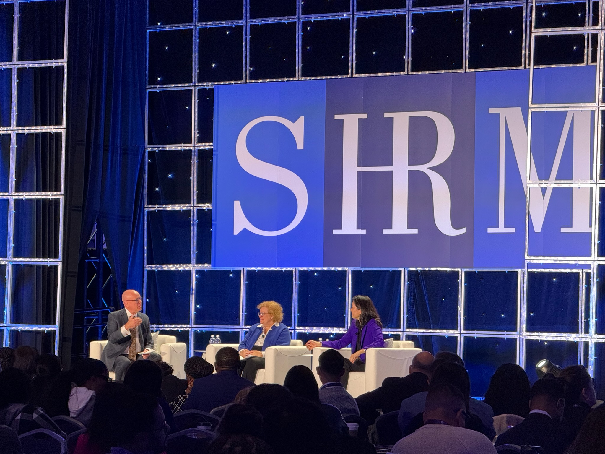 Three panelists engaged in discussion on white chairs at SHRM conference stage with large illuminated SHRM logo backdrop