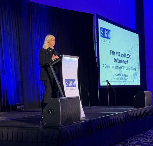 Female speaker in black blazer presenting at podium with SHRM Blueprint branding and Title VII EEOC Enforcement slide