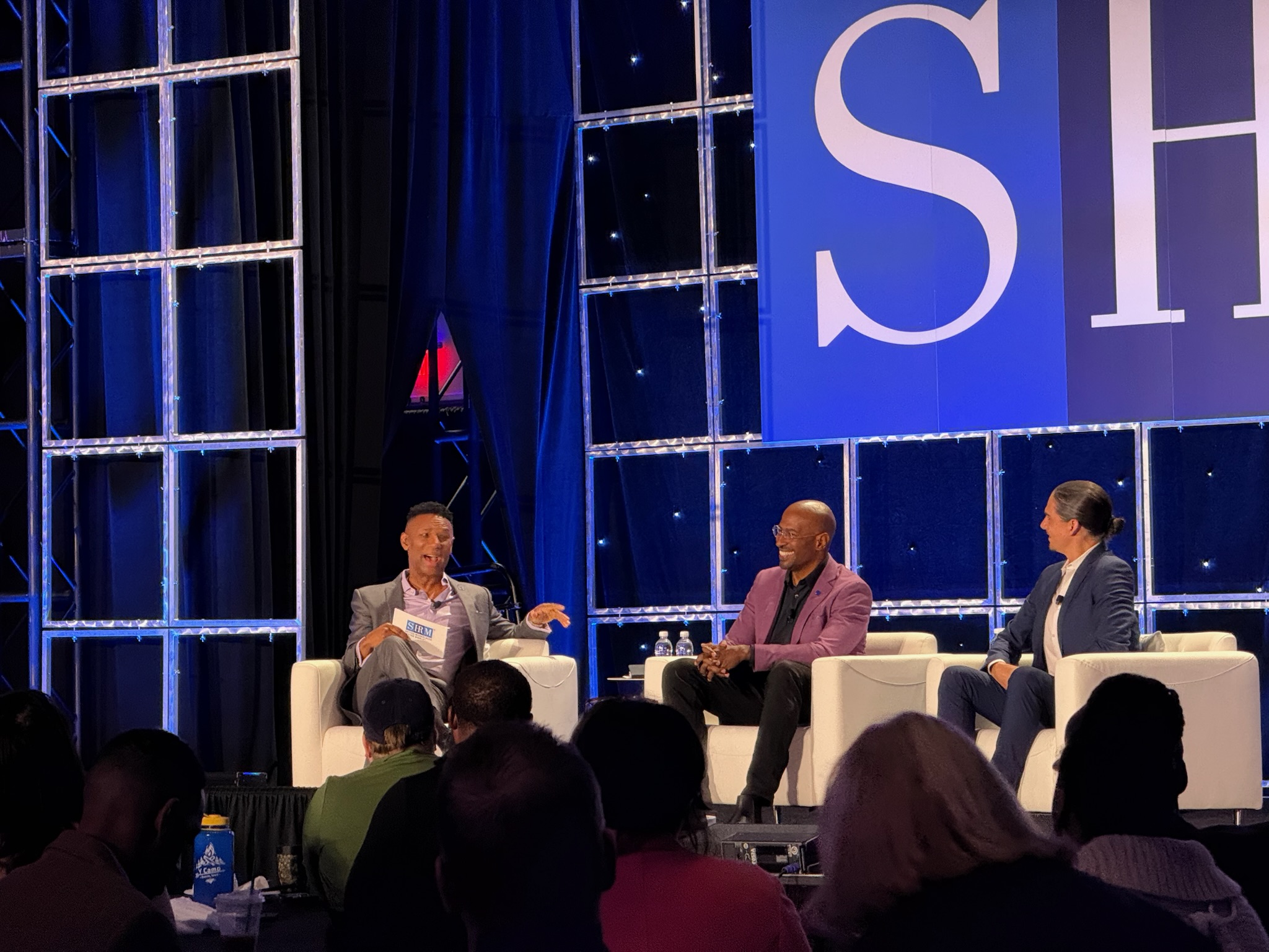Three panelists seated in white chairs on conference stage discussing before audience, with large SHRM branded backdrop