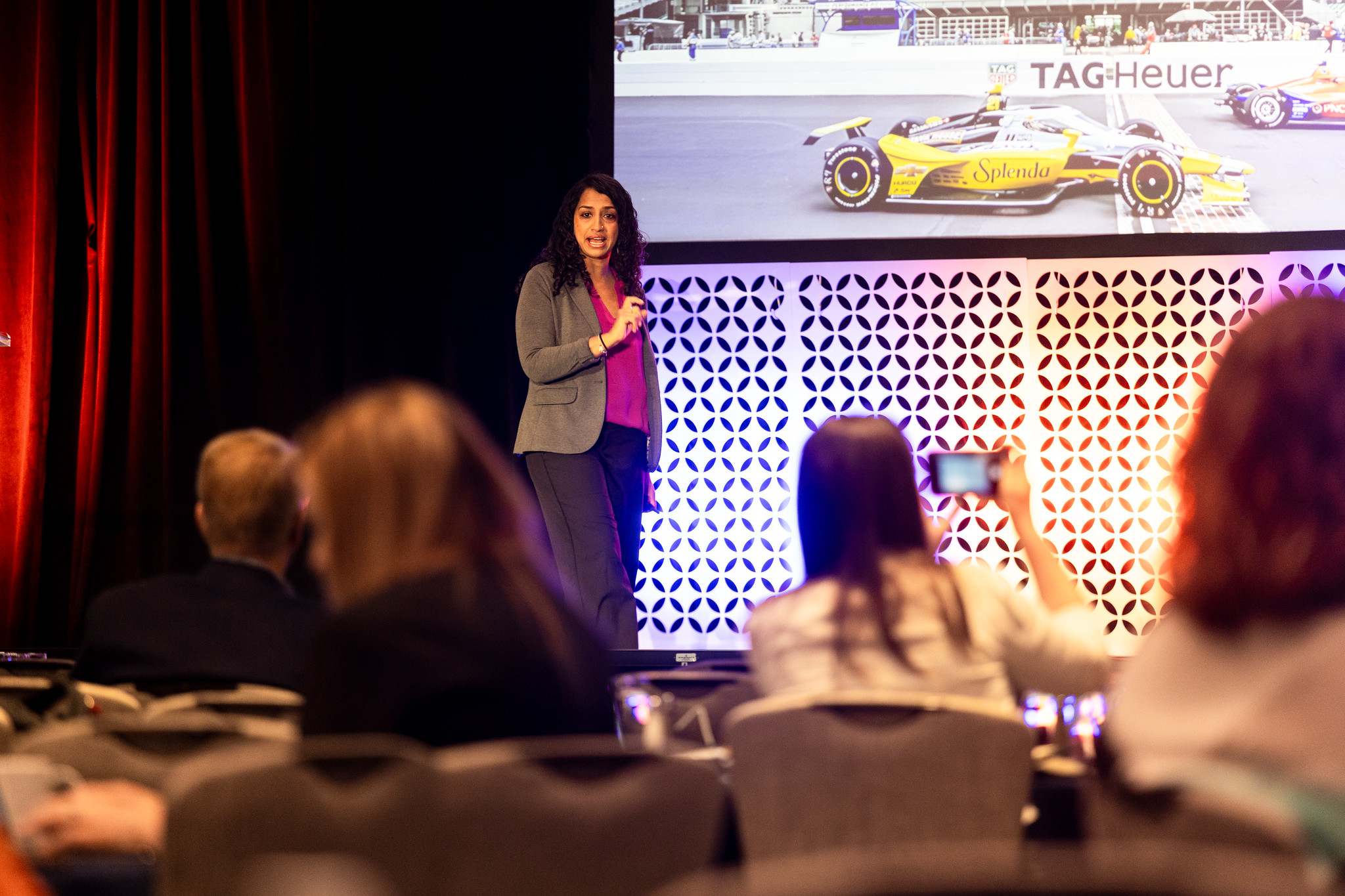 A woman presents on stage during DEAMcon, with indy race cars on the screen behind her, and audience members in front of her.