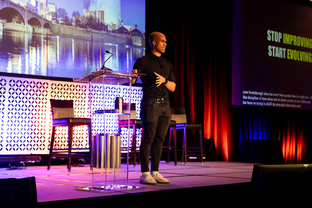 A man stands near a podium on the DEAMcon stage during his keynote presentation.