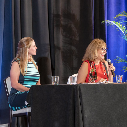 Janet Fiore speaks while Jennifer Polcer listens while both seated behind a table on the stage