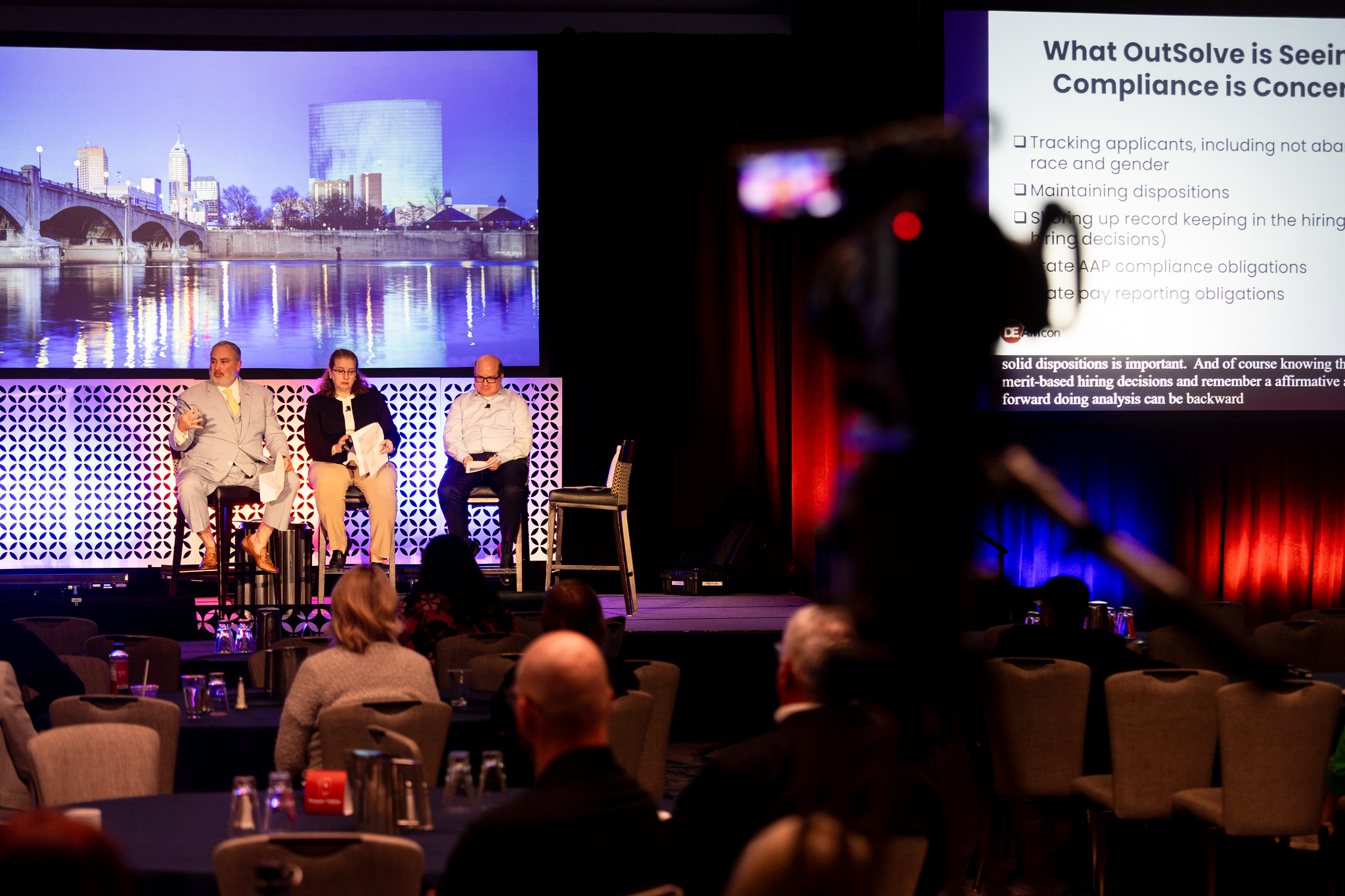 A video camera runs in the foreground, while two men and a woman present on the DEAMcon stage in the distance.