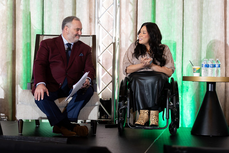 A man sit in a chair on the DEAMcon stage next to a woman in a wheelchair.