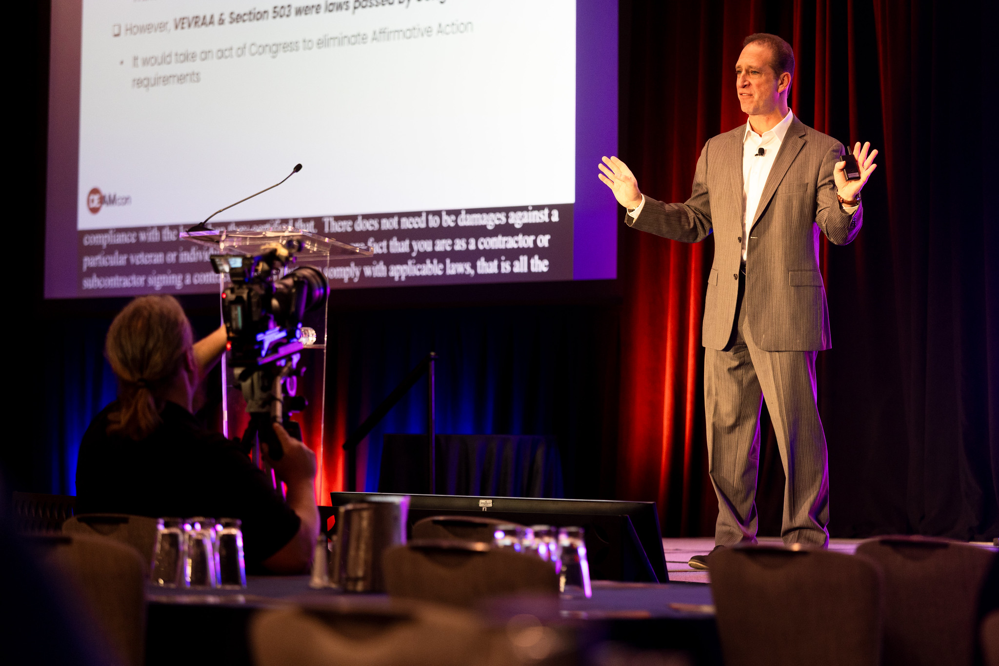 A man presents on the DEAMcon stage near a podium, with a screen behind him displaying presentation slides.