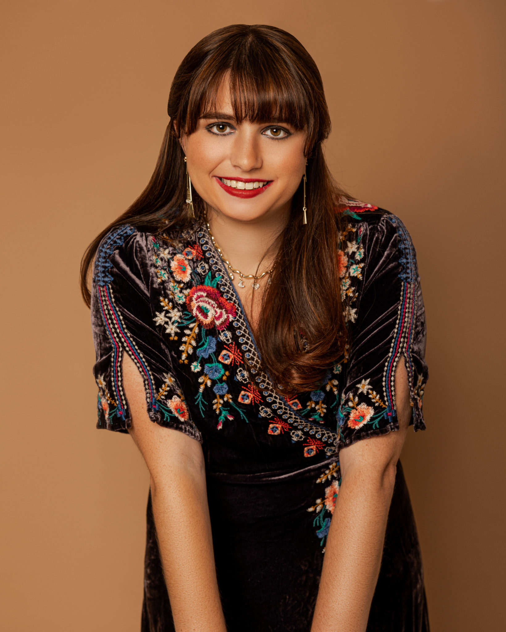 Haley Moss: Pictured is a female in her 20's with dark brown hair, bangs, and a boho-style black dress with colorful embroidering around the neckline