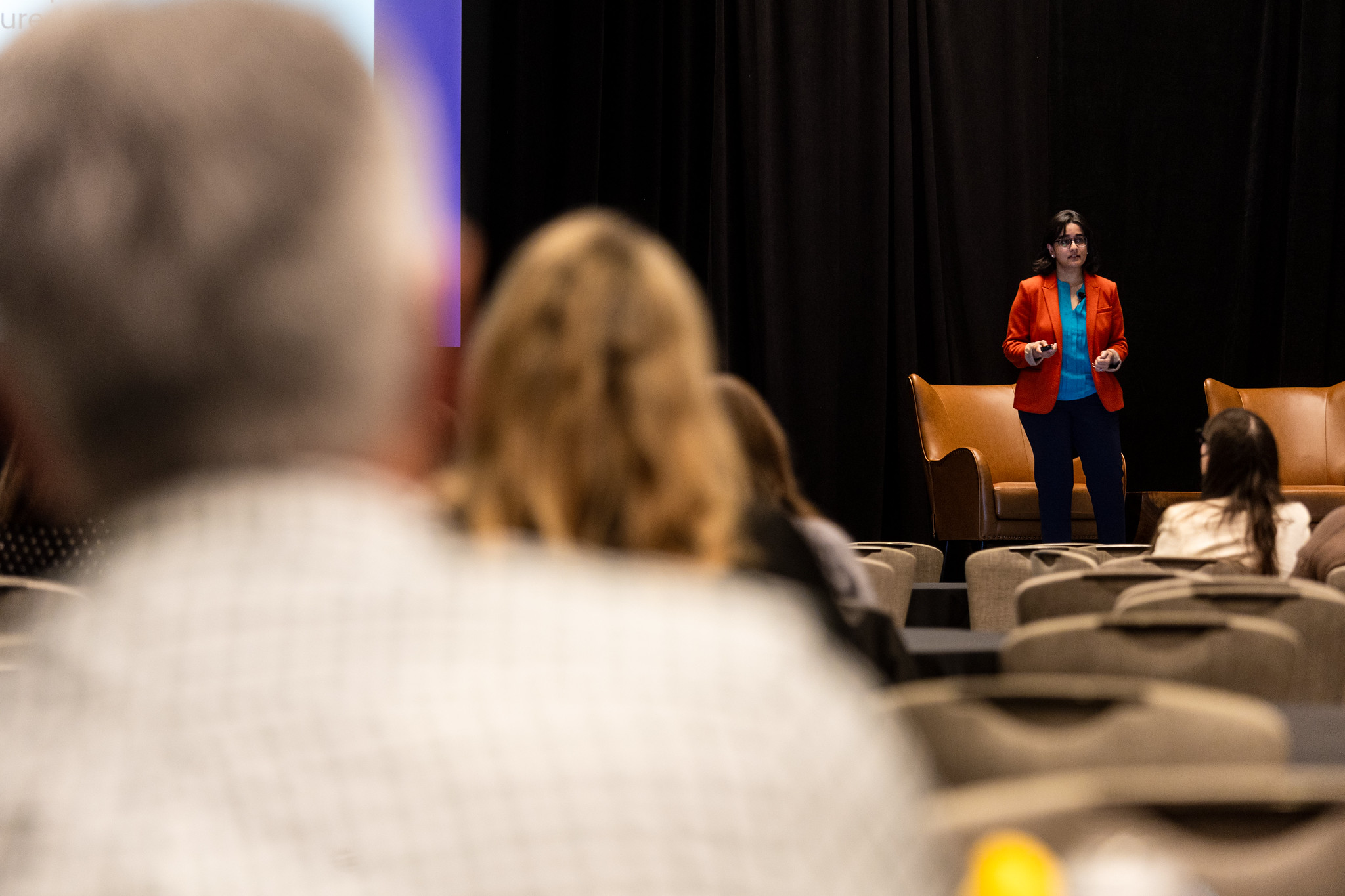 A woman , shown in the background, presents during DEAMcon while attendees look on.