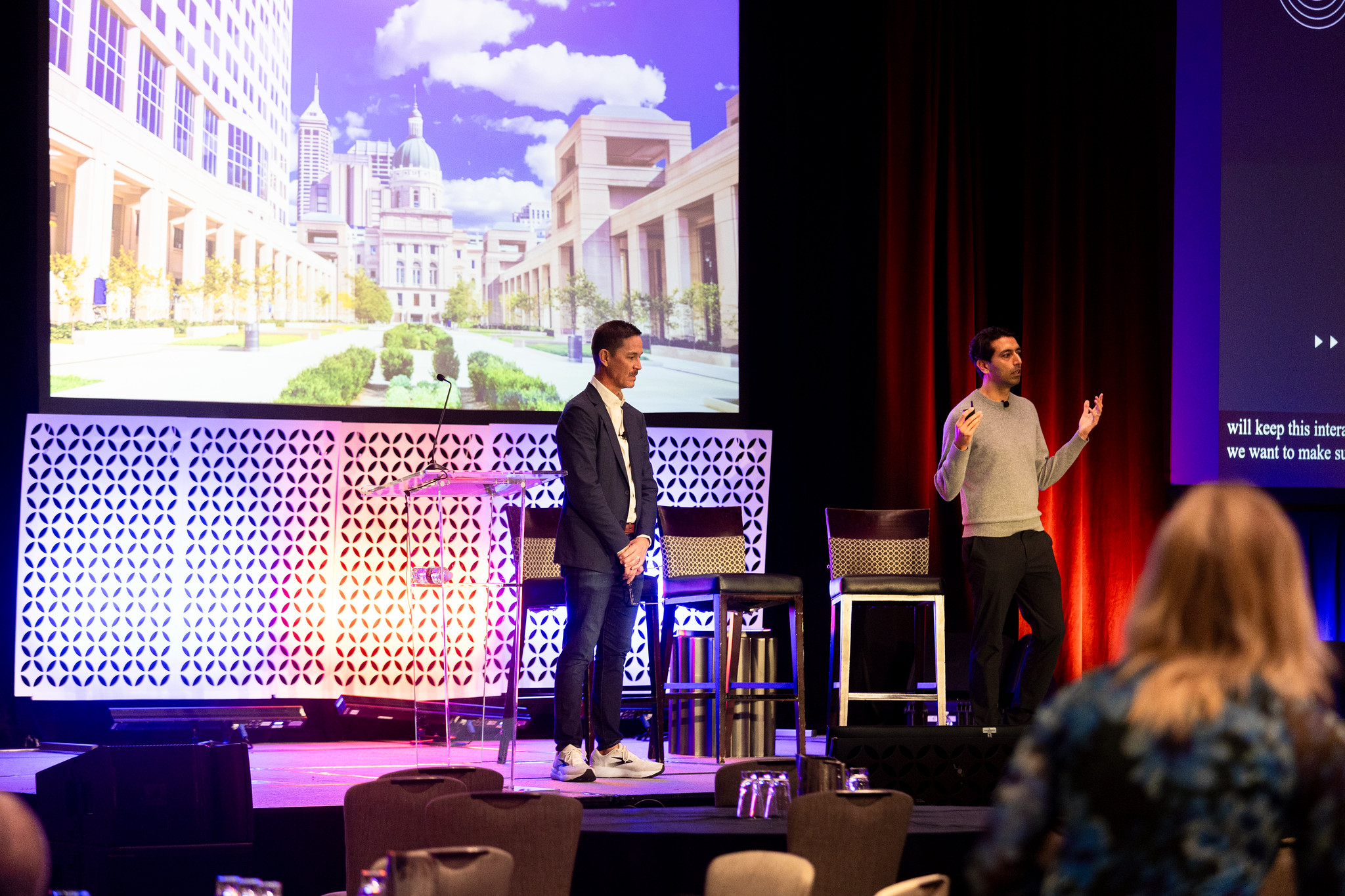 Two men stand on the DEAMcon stage during their presentation with the Indiana State Capital building displayed on the screen in the background.