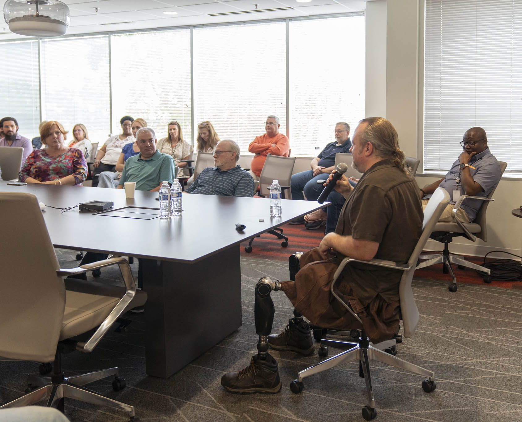 A man with prosthetic legs gives a presentation seated at a conference table, facing an attentive audience seated around and listening actively.