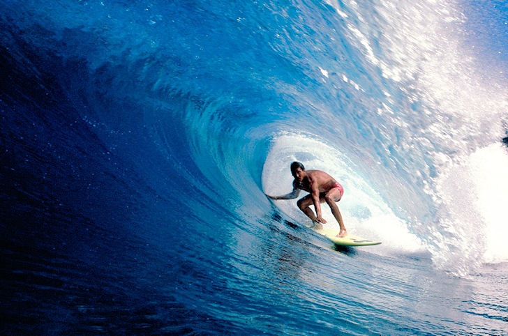 Image of dark blue, rolling waters and surf, with a male surfer on the surfboard at the center